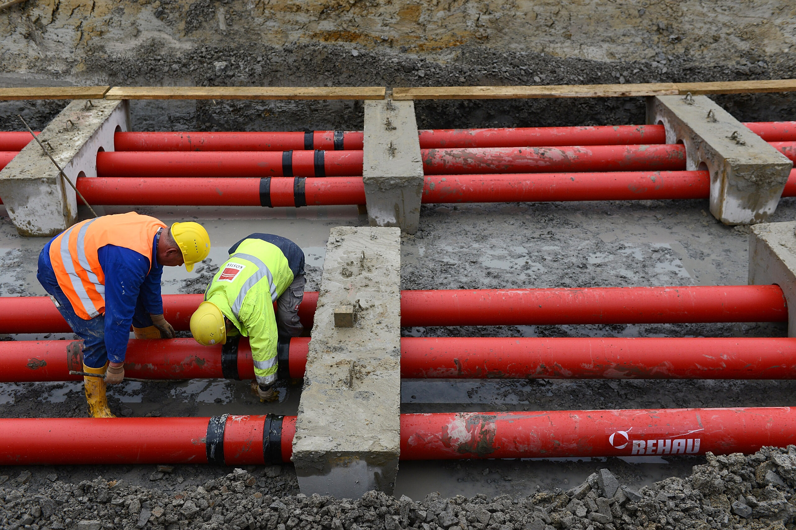 Two workers in yellow helmets and orange and yellow safety vests bend over six red pipes at a construction site.