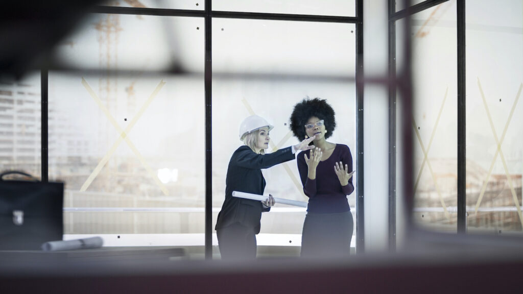 A woman with a hard hat talks to her female colleague on a construction site. They are both in dark suits, in a room with floor-to-ceiling windows.