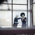 A woman with a hard hat talks to her female colleague on a construction site. They are both in dark suits, in a room with floor-to-ceiling windows.