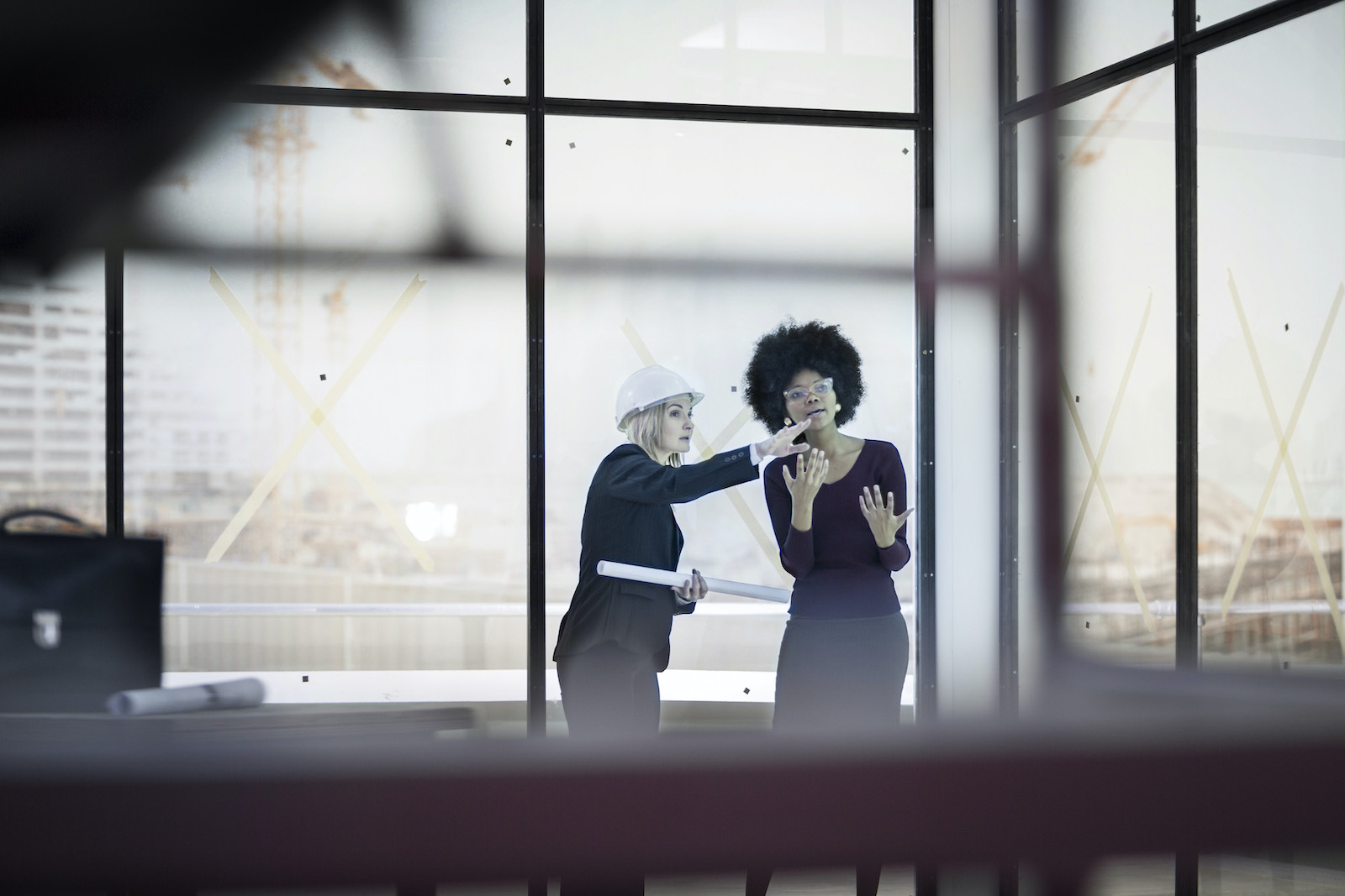 A woman with a hard hat talks to her female colleague on a construction site. They are both in dark suits, in a room with floor-to-ceiling windows.
