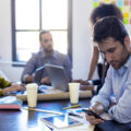 A man in business attire looks at his phone as the rest of his coworkers sit a table with laptops and talk with each other.