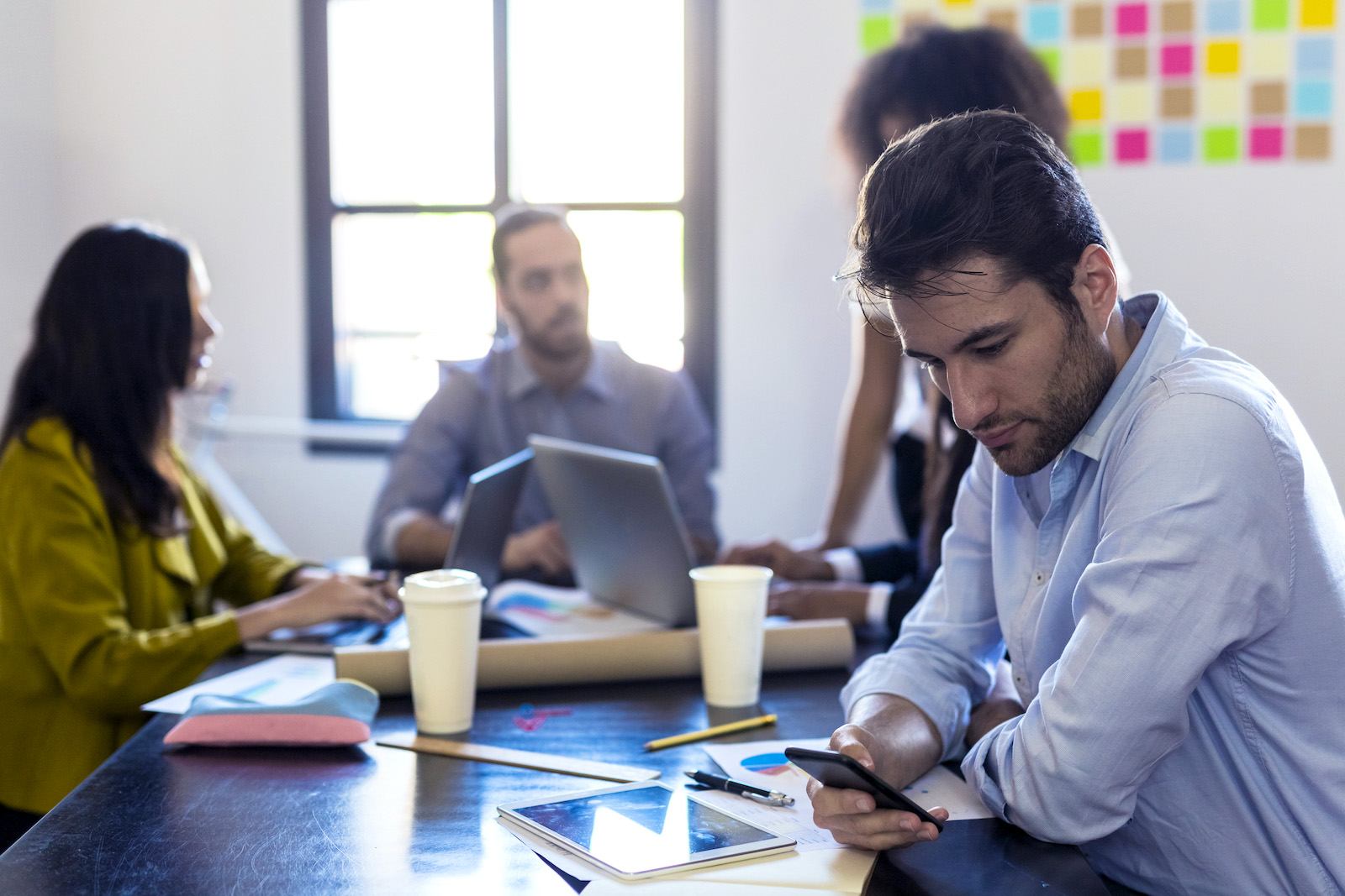 A man in business attire looks at his phone as the rest of his coworkers sit a table with laptops and talk with each other.
