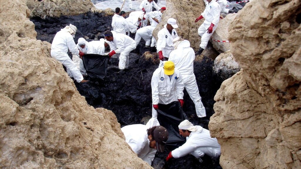A crowd of workers in white protective jumpsuits and hats put plastic over a black oil spill between rocks