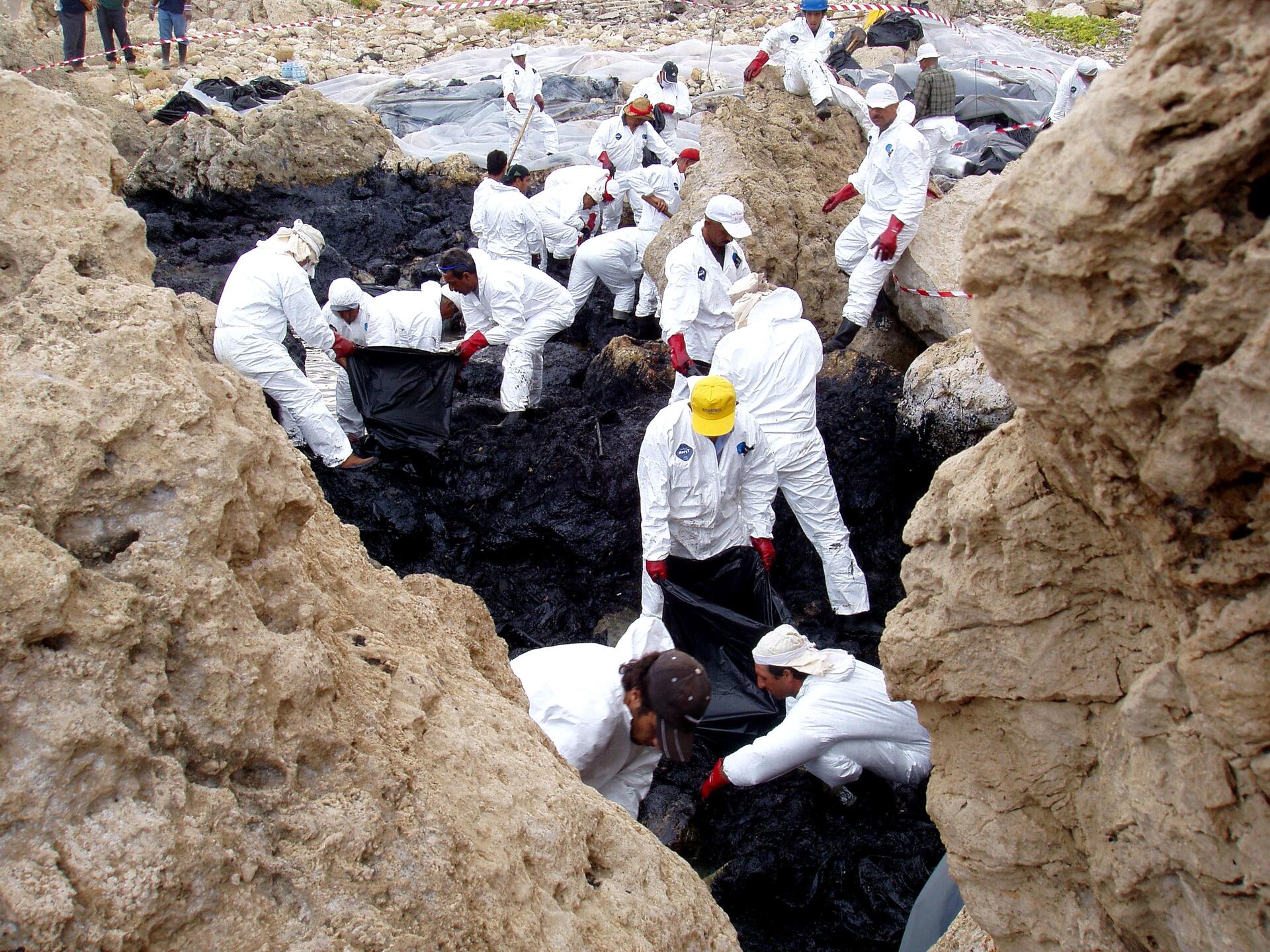 A crowd of workers in white protective jumpsuits and hats put plastic over a black oil spill between rocks