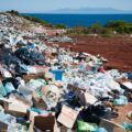 A towering pile of garbage and plastic dwarf a nearby goat. The sea can be seen in the background.