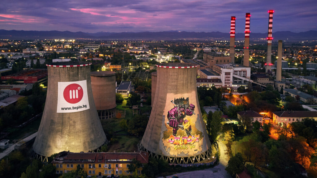 A bird's eye view of a power plant at twilight, with two cement towers in the foreground, and three smokestacks in the background. A city is lit up behind the plant.