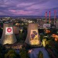 A bird's eye view of a power plant at twilight, with two cement towers in the foreground, and three smokestacks in the background. A city is lit up behind the plant.