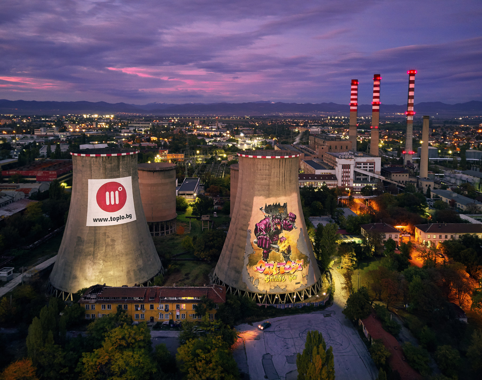A bird's eye view of a power plant at twilight, with two cement towers in the foreground, and three smokestacks in the background. A city is lit up behind the plant.