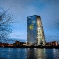 A tall office building on a river, lit up at night with the sign of the Euro.