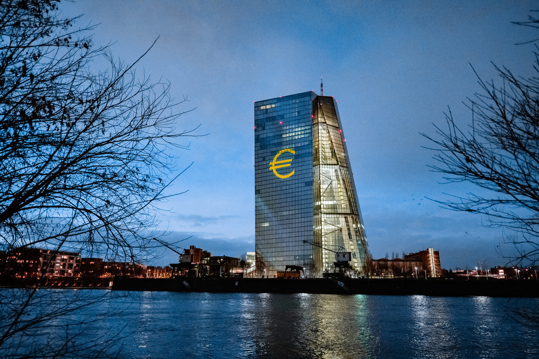 A tall office building on a river, lit up at night with the sign of the Euro.
