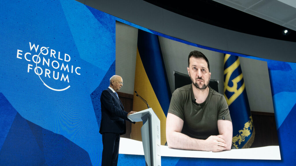 Ukrainian president Zelensky can be seen on a large screen as he calls in virtually to the World Economic Foum. An older man at a podium stands against the virtual screen.