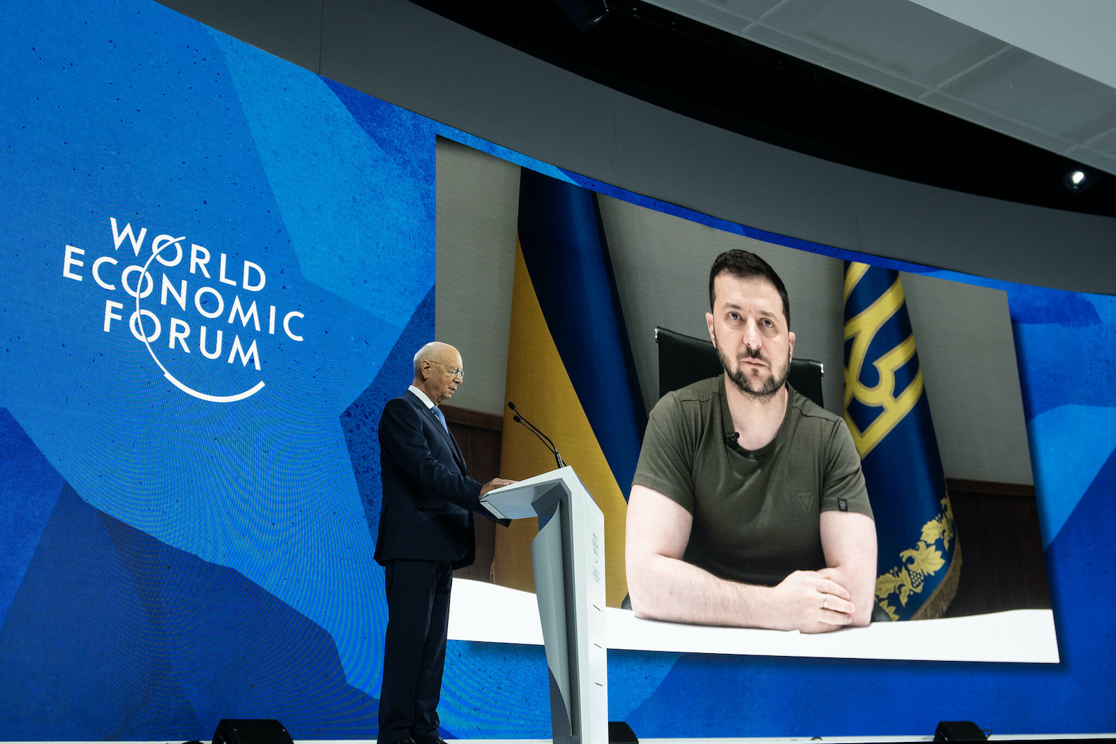 Ukrainian president Zelensky can be seen on a large screen as he calls in virtually to the World Economic Foum. An older man at a podium stands against the virtual screen.