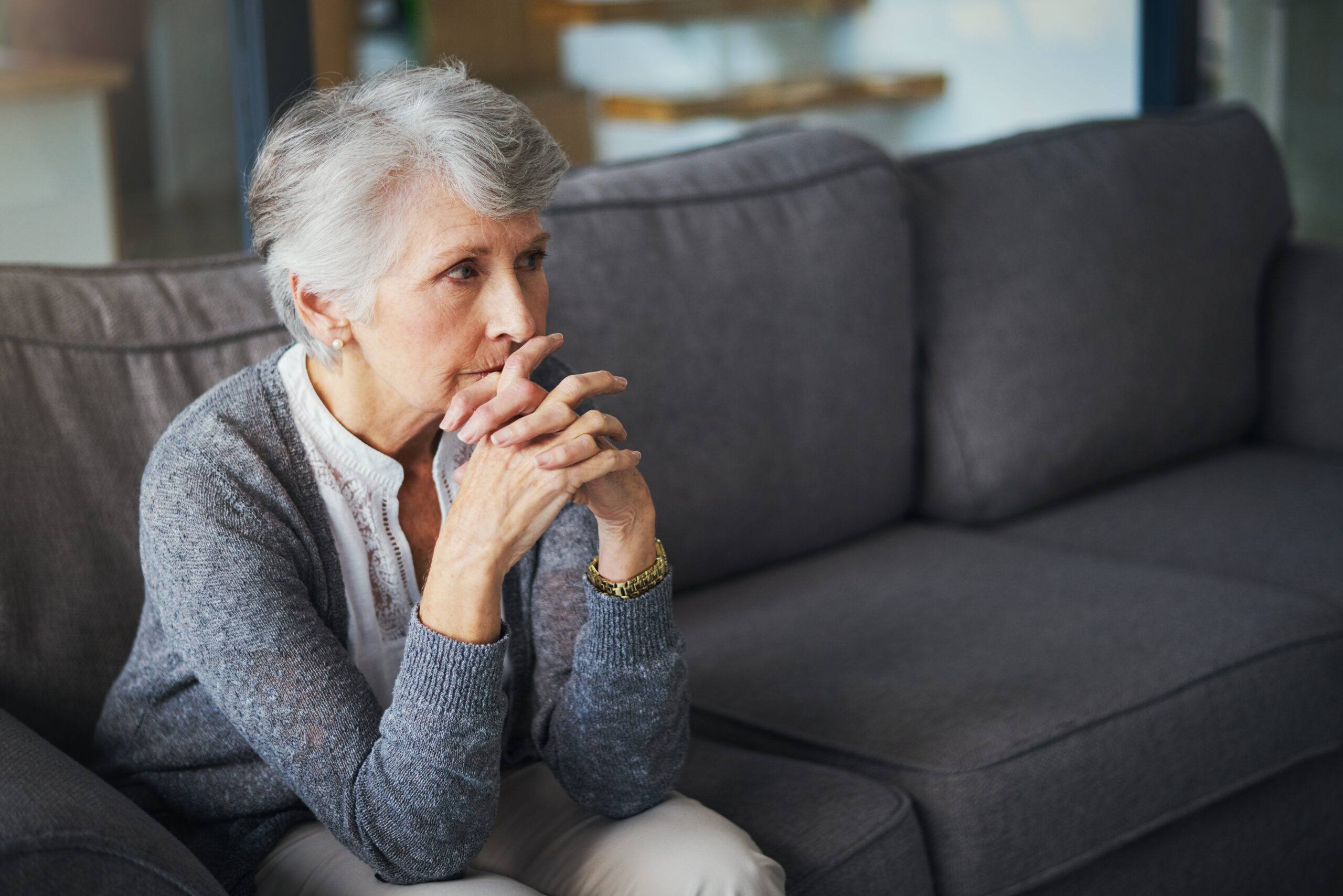 An older woman with grey hair and a cardigan sits on a grey couch with her chin on her folded hands.