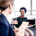 A Black woman in business attire smiles and speaks animatedly to her colleague in an office setting.