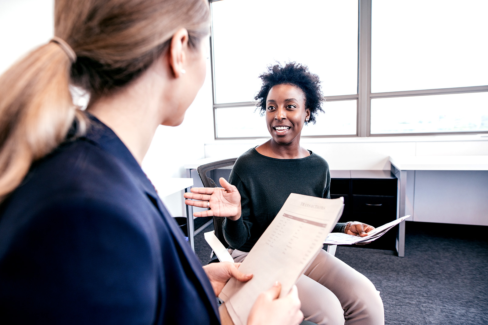 A Black woman in business attire smiles and speaks animatedly to her colleague in an office setting.