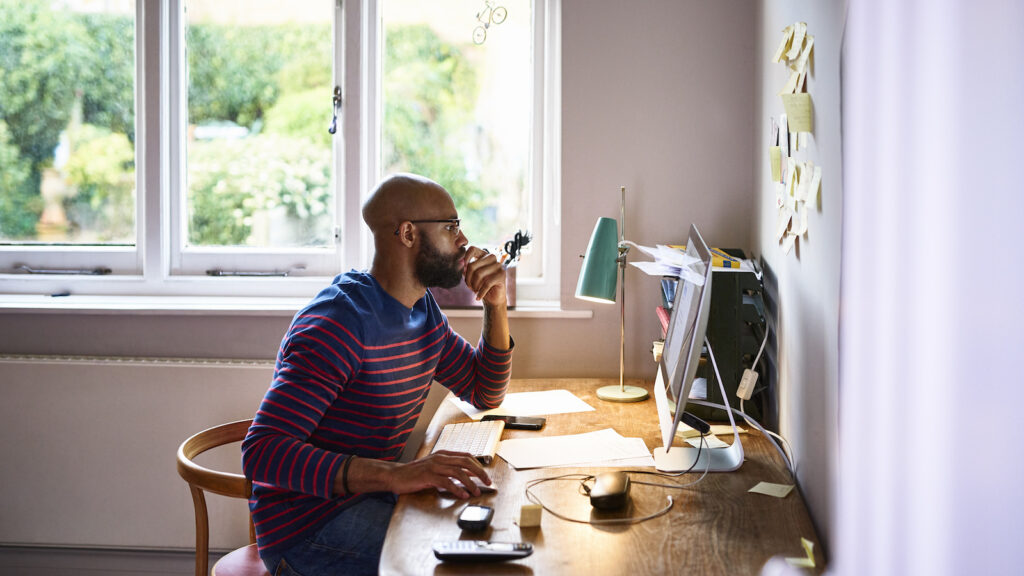 A Black man wearing glasses sits at a desk in his home office, with his chin in his hand as he looks at his computer. A window is behind him, through which green trees can be seen.