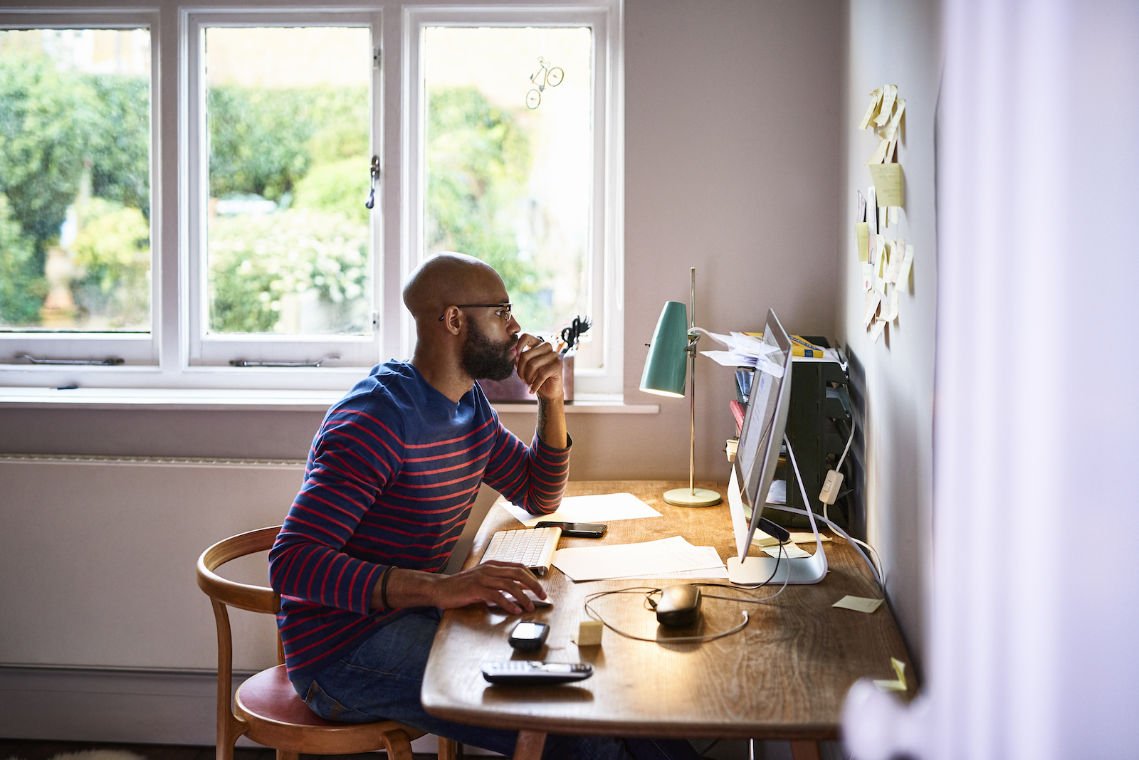 A Black man wearing glasses sits at a desk in his home office, with his chin in his hand as he looks at his computer. A window is behind him, through which green trees can be seen.