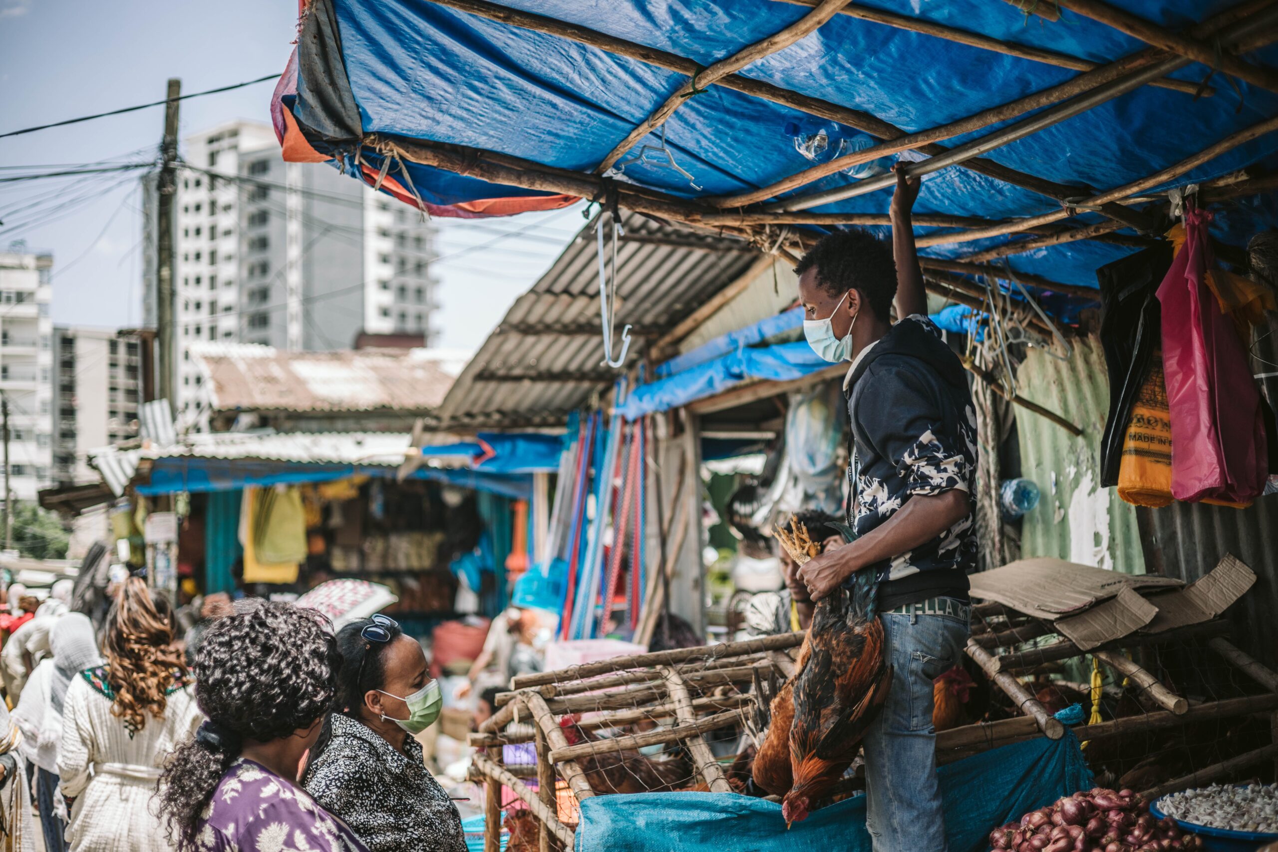 Two women, one in a mask, stand in front of an outdoor market stall. A man wearing a mask stands on a table in the stall, holding poultry.