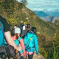 A line of hikers seen from behind walk single file up a green mountain. Blue mountain tops can be seen in the distance