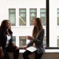 Two women in suits sit on a large window ledge, talking and smiling at each other.