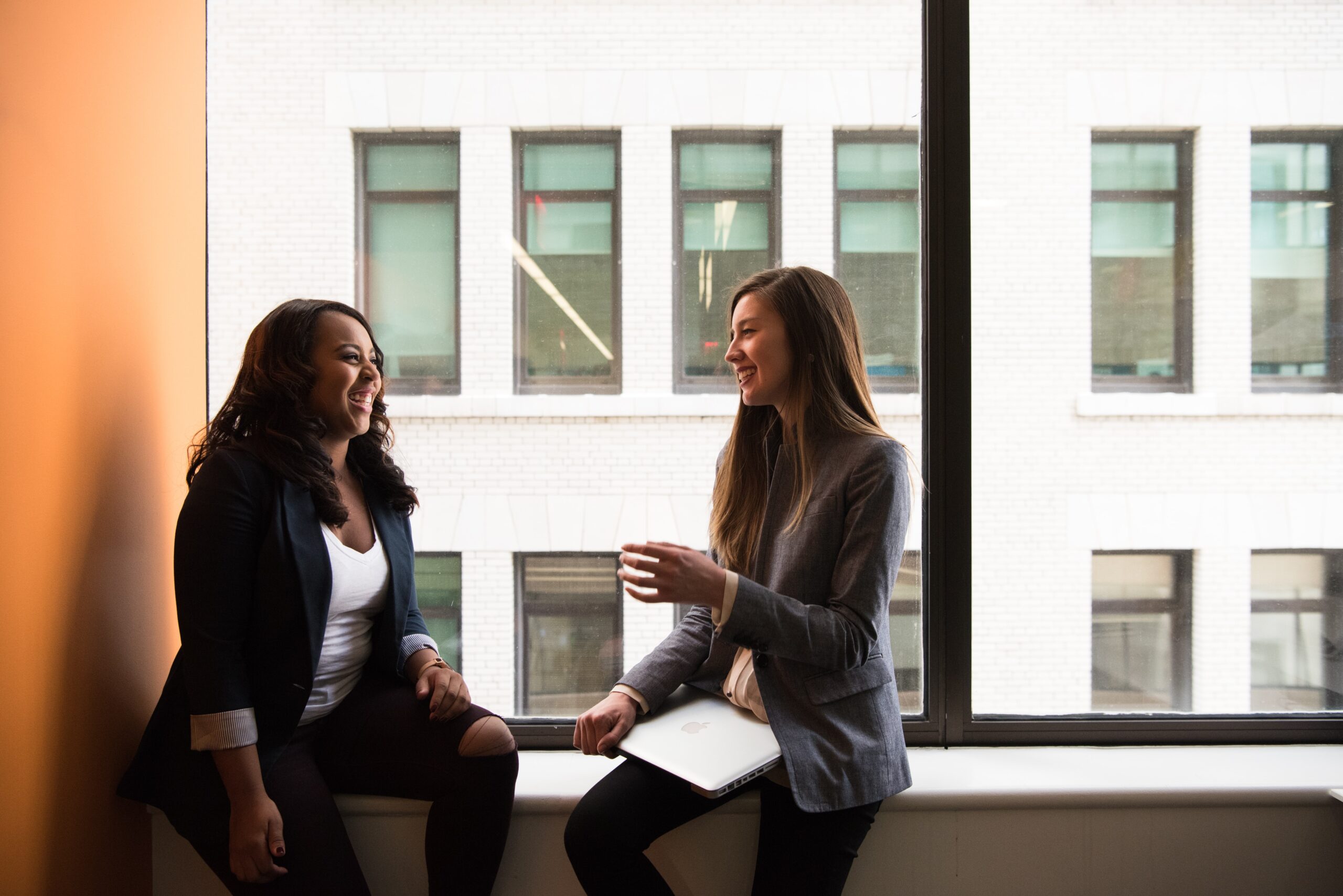 Two women in suits sit on a large window ledge, talking and smiling at each other.
