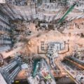 A birds-eye view of a construction site, with tall skyscrapers around a barren middle ground.