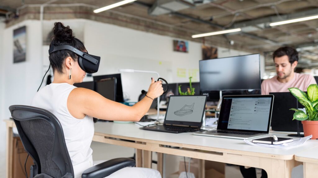 A woman sitting at her office desk plays with a VR headset and hand controls. Her coworker sits opposite her, looking at his computer.