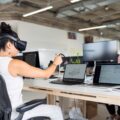 A woman sitting at her office desk plays with a VR headset and hand controls. Her coworker sits opposite her, looking at his computer.