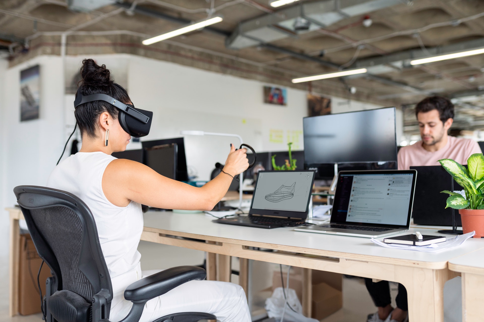 A woman sitting at her office desk plays with a VR headset and hand controls. Her coworker sits opposite her, looking at his computer.