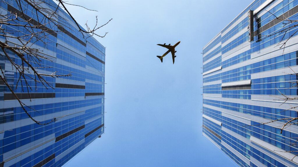 A distant plane seen from below against a blue sky, in between two tall skyscrapers.