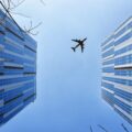 A distant plane seen from below against a blue sky, in between two tall skyscrapers.