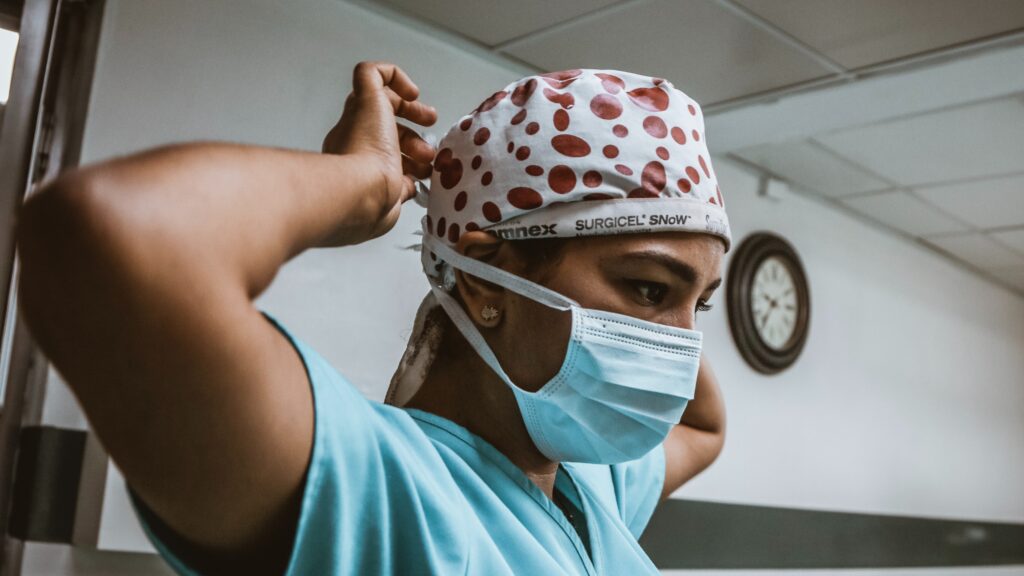 A woman with a serious expression, and wearing scrubs and a surgical cap with red polka dots, ties on her face mask. A white wall and clock can be seen in the background.