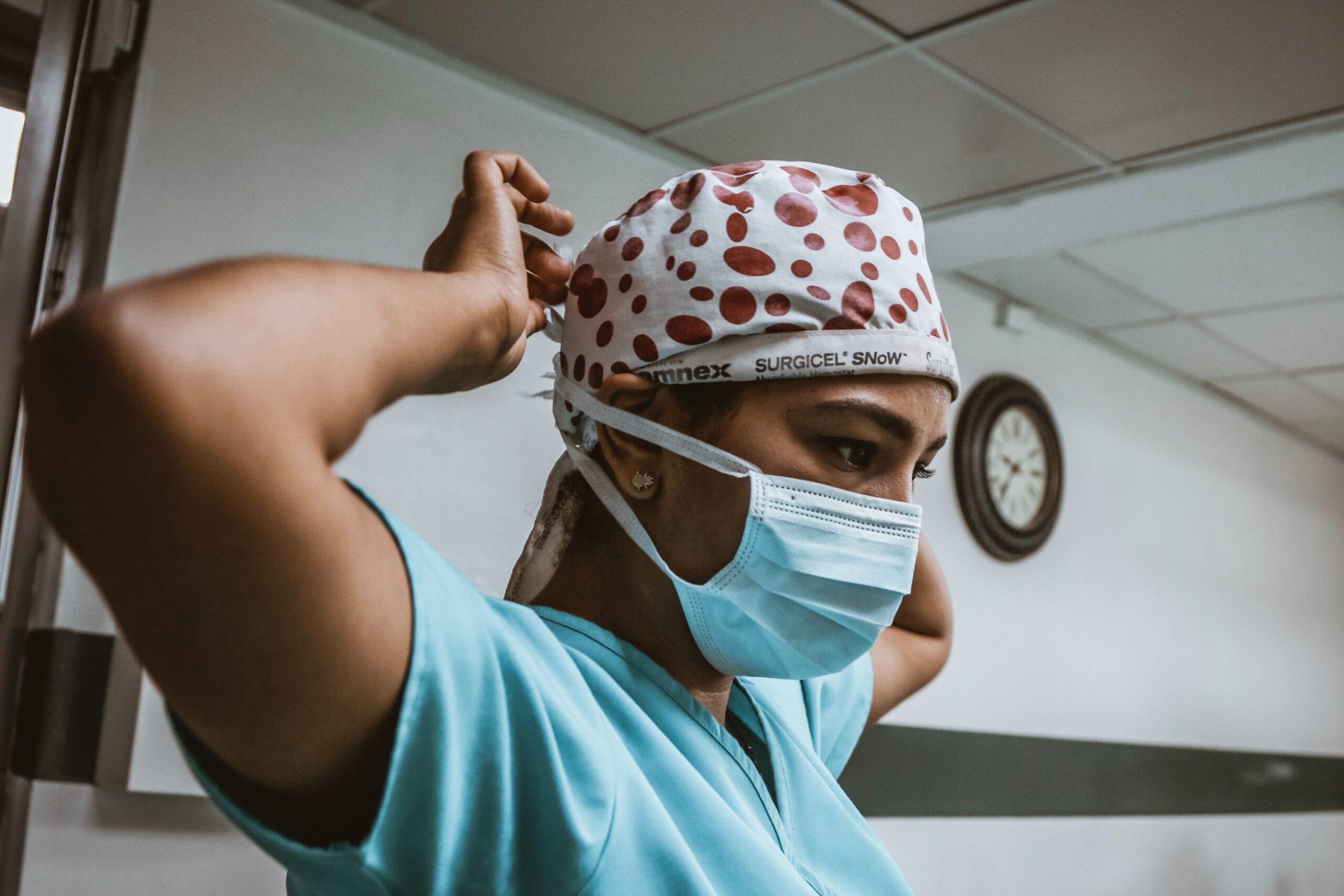 A woman with a serious expression, and wearing scrubs and a surgical cap with red polka dots, ties on her face mask. A white wall and clock can be seen in the background.