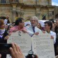 Two men stand next to each other holding up two paper tablets with laws written in spanish. A crowd surrounds them.