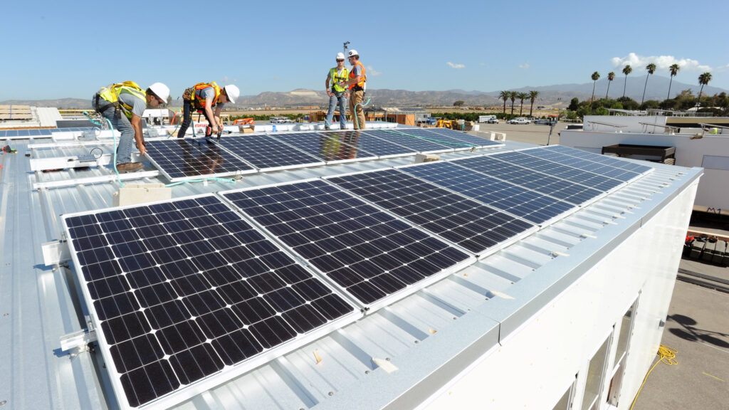 Four people in hard hats and yellow safety vests install solar panels on the roof of a building.