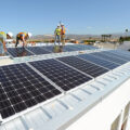 Four people in hard hats and yellow safety vests install solar panels on the roof of a building.