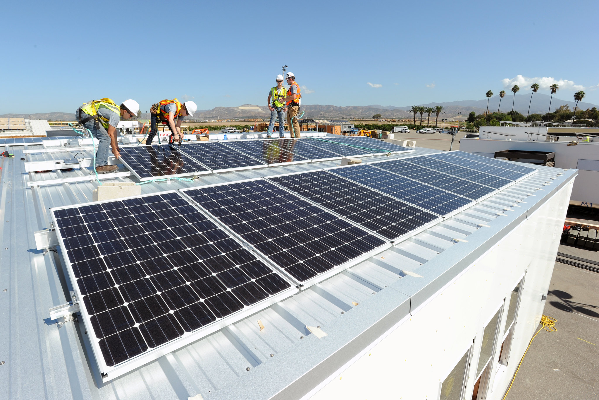 Four people in hard hats and yellow safety vests install solar panels on the roof of a building.
