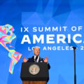 Biden stands at a lectern with the seal of the United States, holding both hands up, palms facing outwards. There is a blue screen behind him with a logo saying 