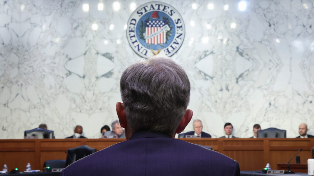 The back of a man's head and shoulders is the foreground. The man is seated and wearing a blue suit jacket. In the background is a raised dais with people seated. The seal of the United States hangs above them on a marble wall.