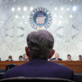 The back of a man's head and shoulders is the foreground. The man is seated and wearing a blue suit jacket. In the background is a raised dais with people seated. The seal of the United States hangs above them on a marble wall.