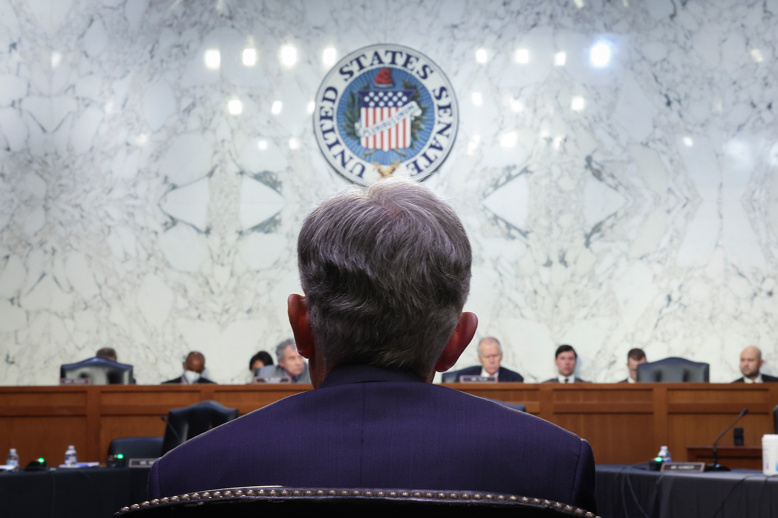 The back of a man's head and shoulders is the foreground. The man is seated and wearing a blue suit jacket. In the background is a raised dais with people seated. The seal of the United States hangs above them on a marble wall.