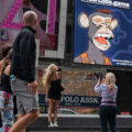 A illustration of a yawning monkey wearing a small bowler hat and orange overalls appears on a billboard over a store. One woman poses beneath it while another woman takes her picture. A man and a woman look up at it as they walk by.