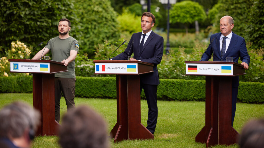 Three men stand behind podiums on a grass lawn. At the far left is Ukrainian President Zelenskyy wearing a green tshirt with the Ukrainian flag on the sleeve. To his right are French President Emmanuel Macron and German Chancellor Olaf Scholz, both in dark suits.