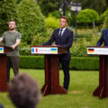 Three men stand behind podiums on a grass lawn. At the far left is Ukrainian President Zelenskyy wearing a green tshirt with the Ukrainian flag on the sleeve. To his right are French President Emmanuel Macron and German Chancellor Olaf Scholz, both in dark suits.