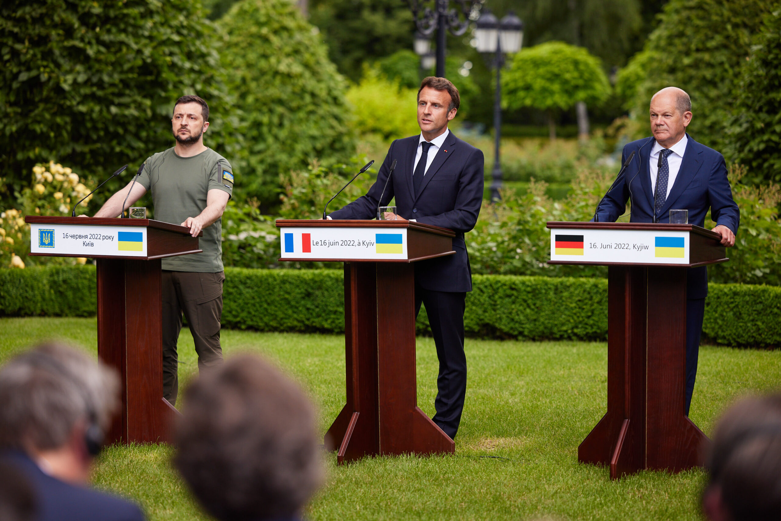 Three men stand behind podiums on a grass lawn. At the far left is Ukrainian President Zelenskyy wearing a green tshirt with the Ukrainian flag on the sleeve. To his right are French President Emmanuel Macron and German Chancellor Olaf Scholz, both in dark suits.