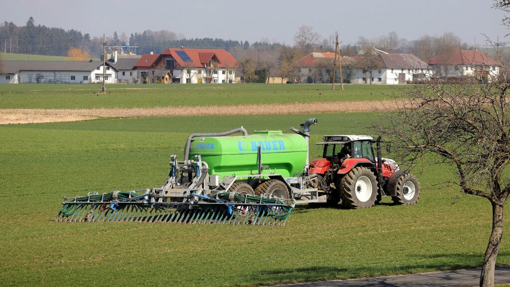 A red tractor pulling a bright green fertilizer tank behind it rolls across a green field. A house with a red tile roof can be seen in the distance.