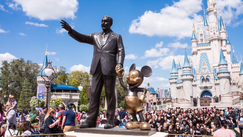 A photo of crowds at Disneyland on a sunny day. A statue of Walt Disney holding Mickey Mouse's hand is in the foreground, with the Magic Castle in the background.