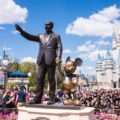 A photo of crowds at Disneyland on a sunny day. A statue of Walt Disney holding Mickey Mouse's hand is in the foreground, with the Magic Castle in the background.
