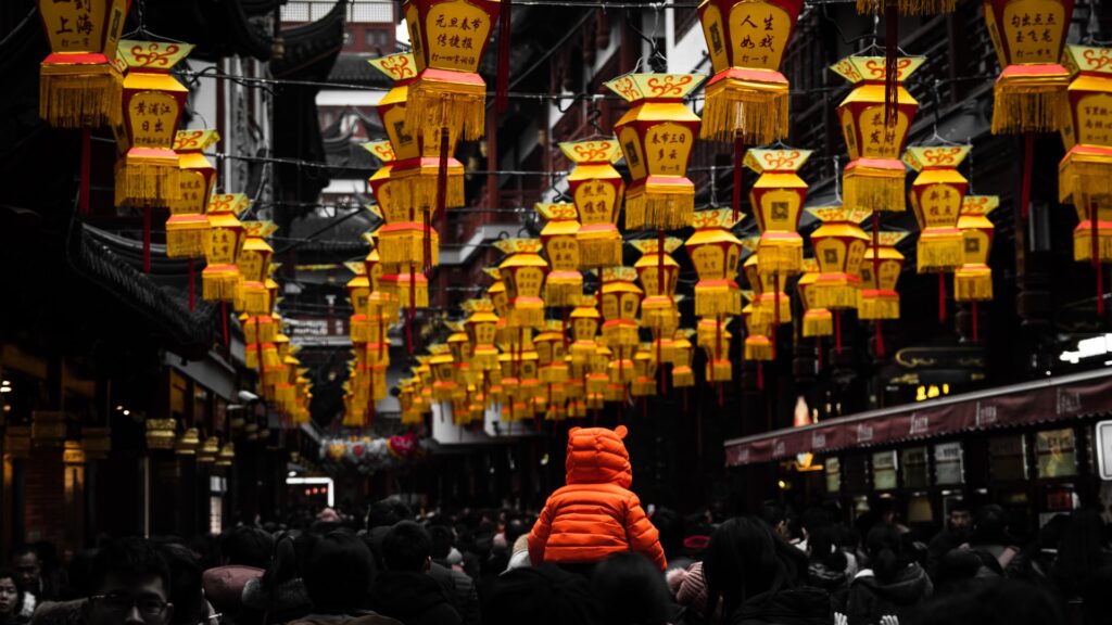 A small child in an orange coat with a hood with ears sits on an adults shoulder in a busy market. The picture is mostly dark except for the rows of bright yellow and red Chinese lanterns hanging from the ceiling.