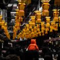 A small child in an orange coat with a hood with ears sits on an adults shoulder in a busy market. The picture is mostly dark except for the rows of bright yellow and red Chinese lanterns hanging from the ceiling.